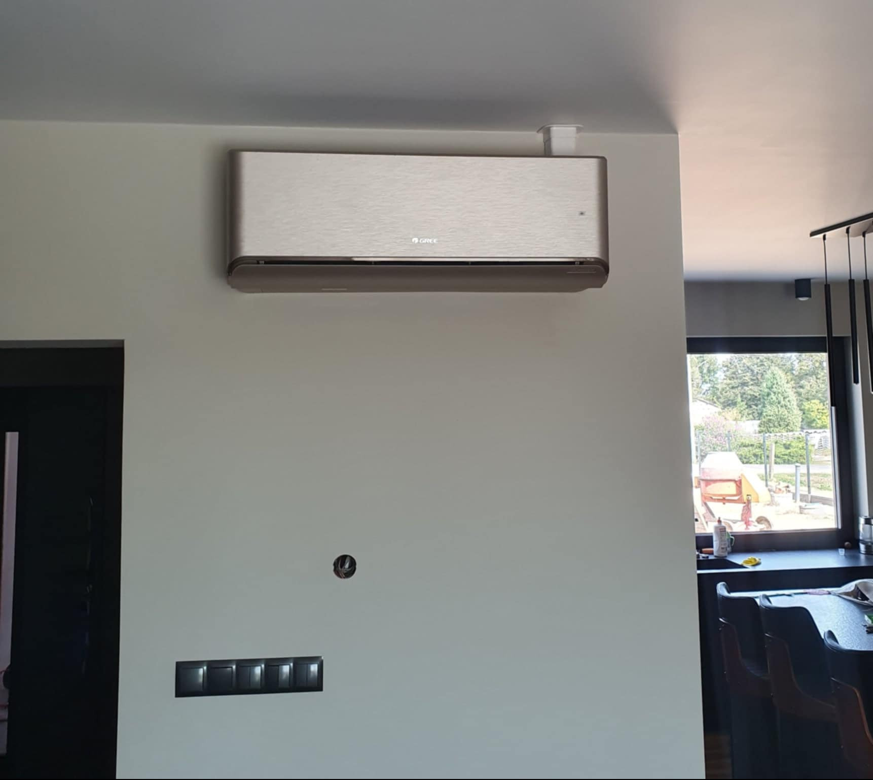 Zamontowany klimatyzator Gree Airy w nowoczesnym wnętrzu, widoczne sterowanie na ścianie i fragment okna z widokiem na ogród.