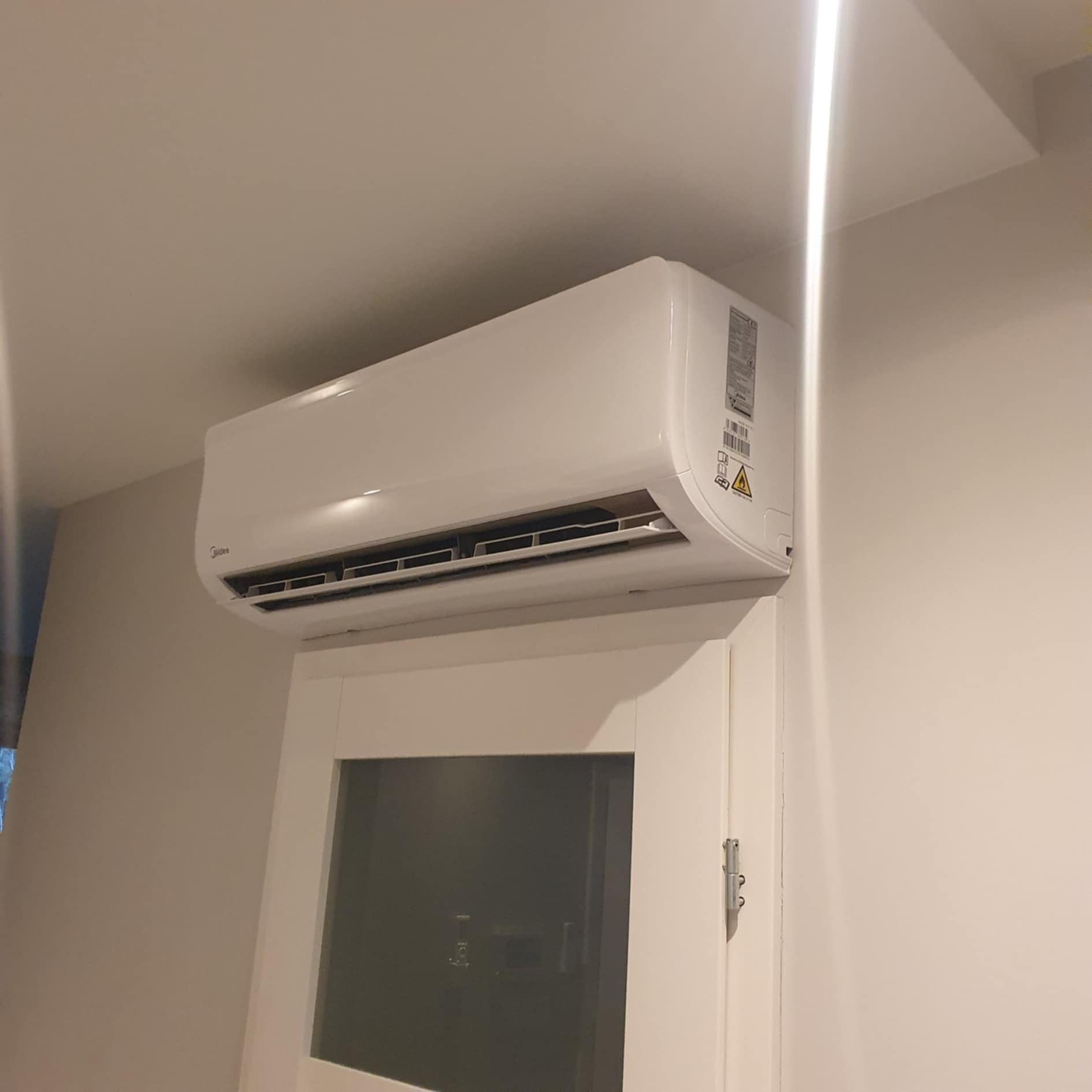 Biała jednostka klimatyzacji Midea zamontowana nad drzwiami w jasnym pomieszczeniu.