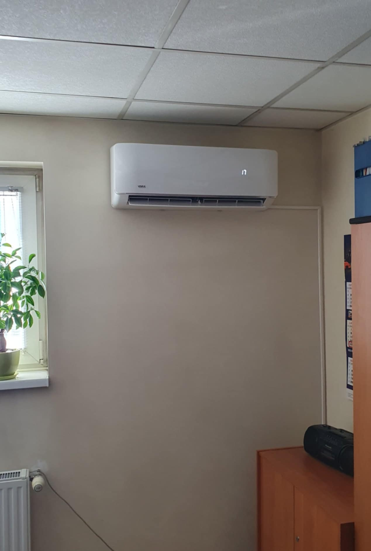 Biały klimatyzator ścienny marki Vivax z wyświetlaczem ustawionym na 17 stopni, zainstalowany w biurze z kasetonowym sufitem, obok okna z zieloną rośliną doniczkową i szafy z kalendarzem.