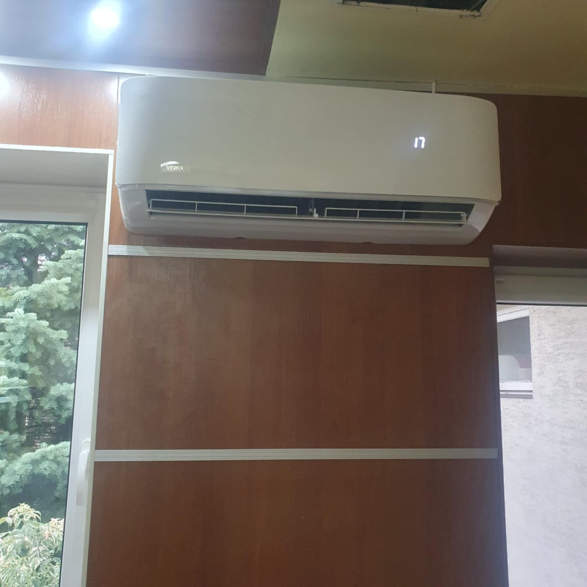 Biała klimatyzacja marki Vivax zamontowana na ścianie z boazerii, obok okna z widokiem na zieleń.