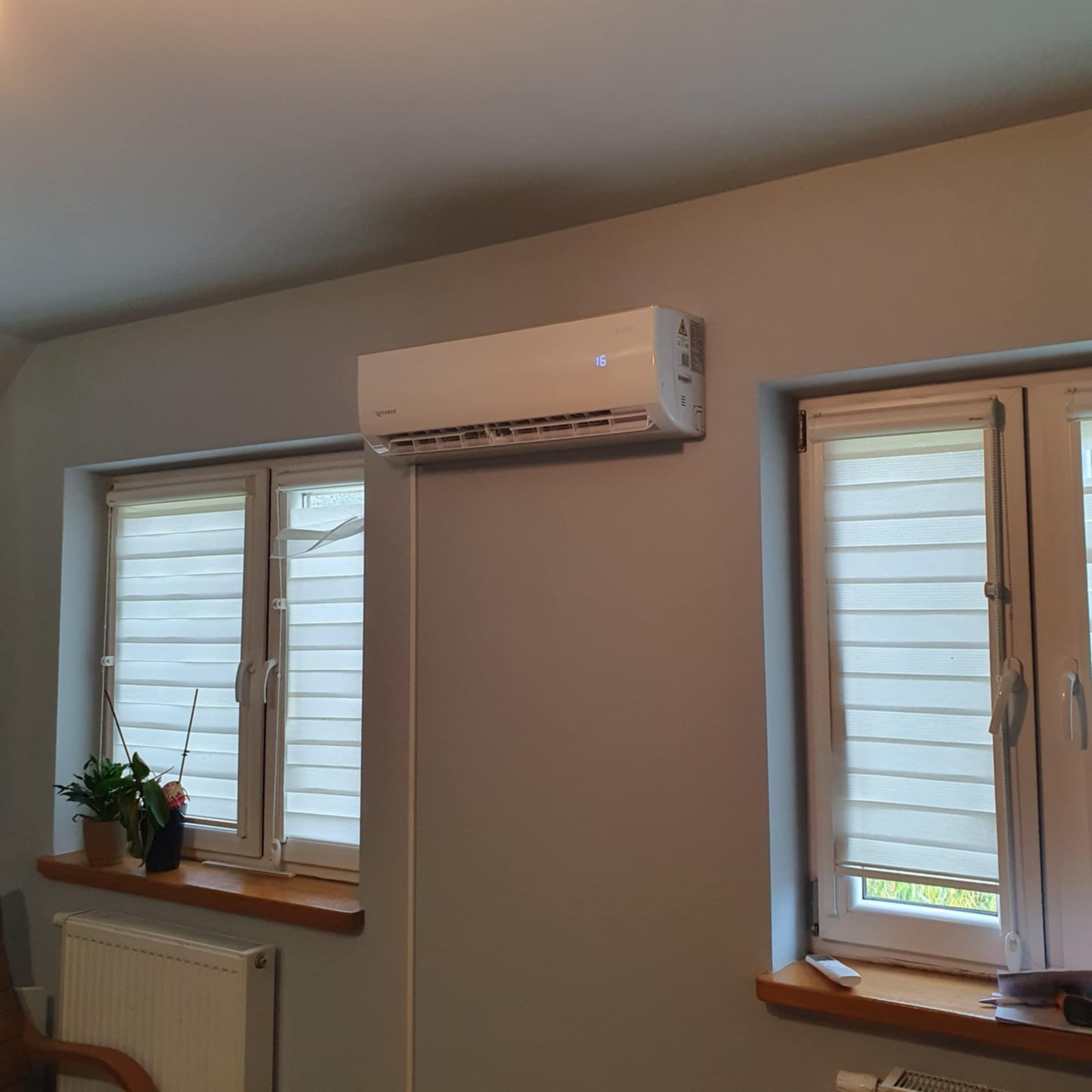 Biały klimatyzator ścienny zamontowany w pokoju z dwoma oknami z żaluzjami poziomymi, widoczny przewód zasilający wzdłuż ściany.