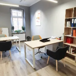 Jasne, przestronne biuro z dwoma biurkami, szarą ścianą i regałem z książkami i segregatorami; roślina doniczkowa w prawym dolnym rogu.