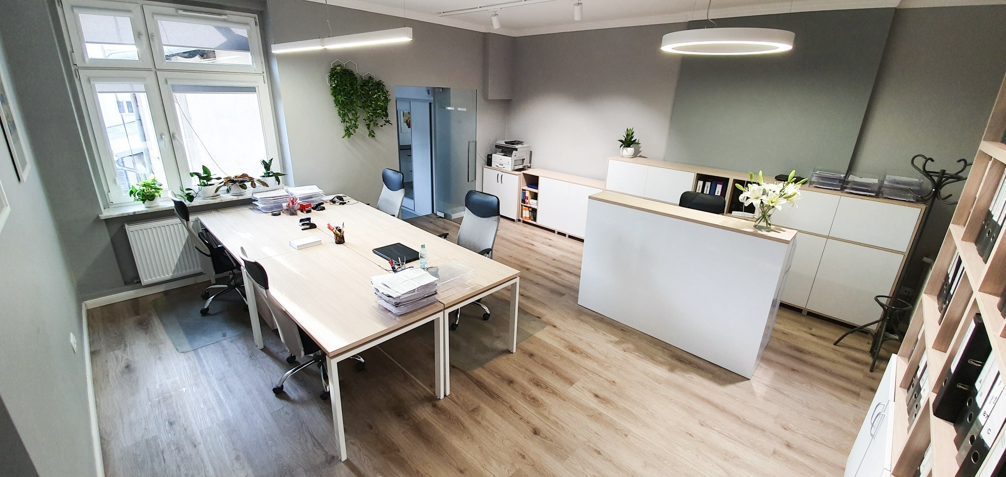 Jasne biuro z recepcją, biurkami z dokumentami, roślinami doniczkowymi i nowoczesnym oświetleniem.