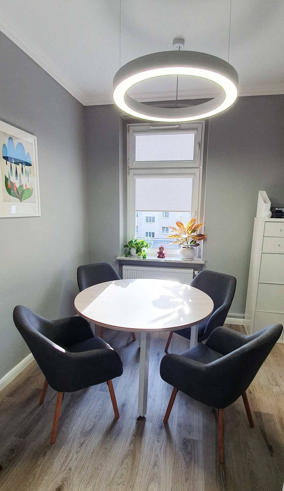 Jasne biuro z okrągłym stołem, czterema krzesłami z ciemnej tkaniny na drewnianych nogach, oświetlone nowoczesną okrągłą lampą wiszącą; na parapecie rośliny doniczkowe i mała figurka.
