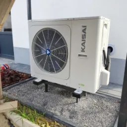 Zewnętrzna jednostka pompy ciepła Kaisai zamontowana na metalowej konstrukcji przy ścianie budynku, otoczona szarym żwirem i elementami ogrodu z piłką w tle.