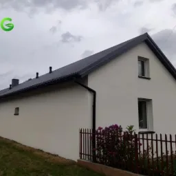 Dom jednorodzinny z czarną blachodachówką, białymi ścianami i ciemnymi rynnami, widoczny z boku, z fragmentem czerwonego ogrodzenia i fioletowymi kwiatami na pierwszym planie, pochmurne niebo.