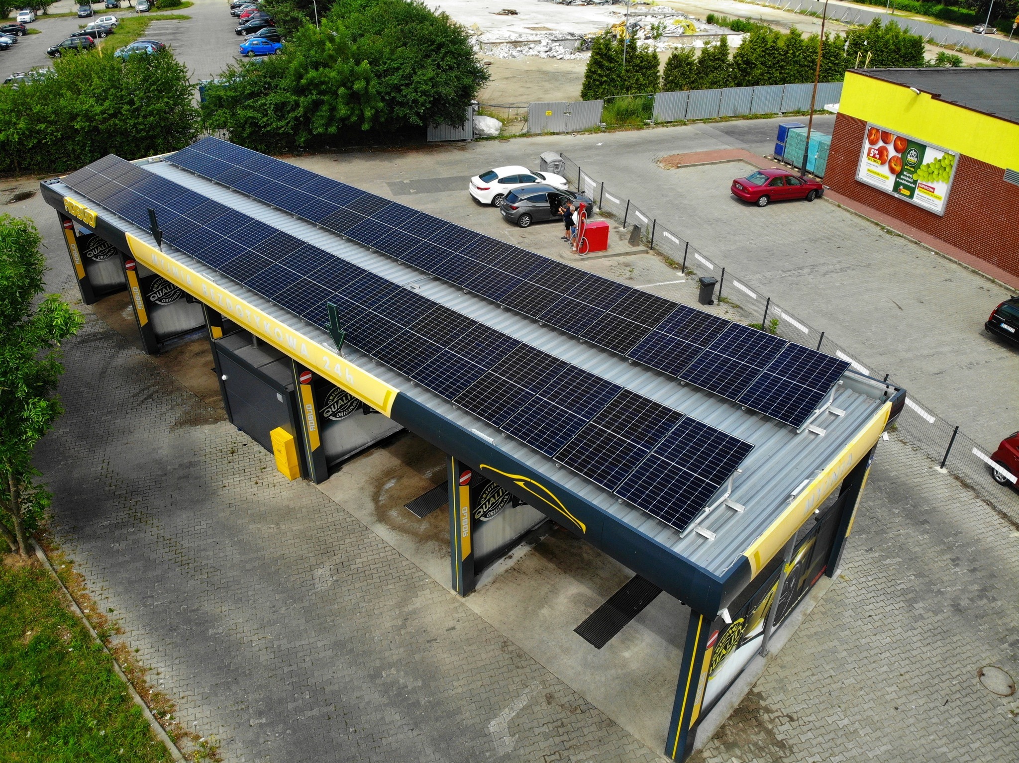 Widok z góry na myjnię samochodową z panelami słonecznymi na dachu, obok zaparkowane samochody i budynek sklepu spożywczego z reklamą owoców.