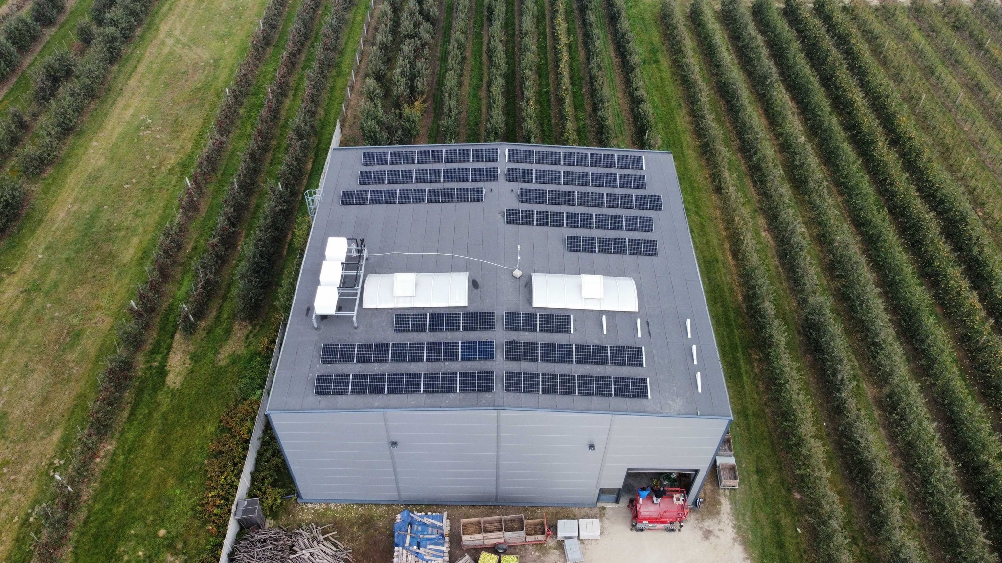 Widok z drona na magazyn z panelami słonecznymi na dachu, otoczony sadem owocowym, z widocznym sprzętem rolniczym i skrzyniami na owoce przy wejściu.