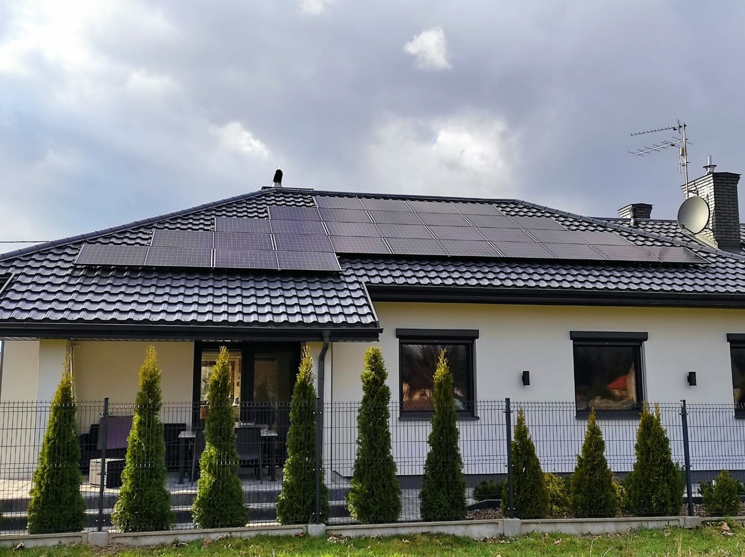 Dom jednorodzinny z panelami słonecznymi na dachu pokrytym czarną dachówką, widoczne również okna z roletami zewnętrznymi, metalowe ogrodzenie i rząd tuj.
