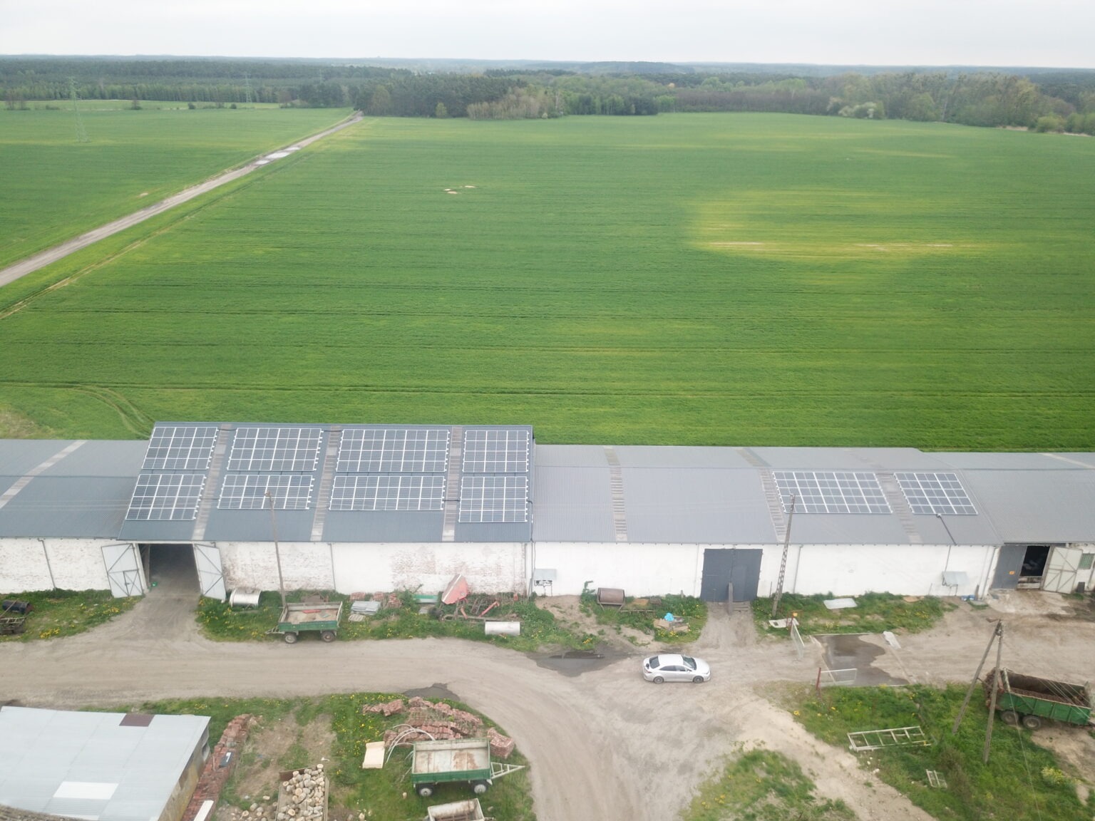 Widok z lotu ptaka na budynek gospodarczy w Międzyrzeczu z zamontowanymi panelami słonecznymi na dachu, otoczony polami i lasami. Na podwórzu widoczny samochód osobowy i przyczepy.