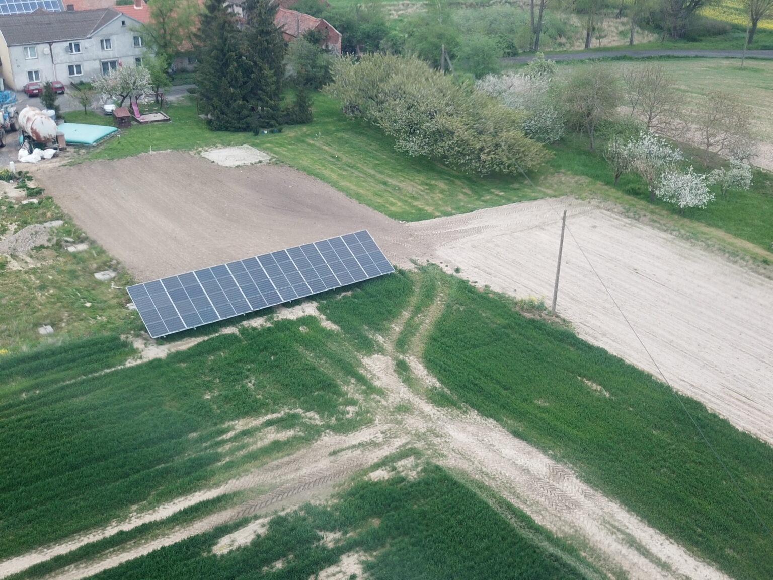 Widok z góry na zainstalowane panele słoneczne na polu, widoczne ślady opon, w tle budynki mieszkalne oraz drzewa.