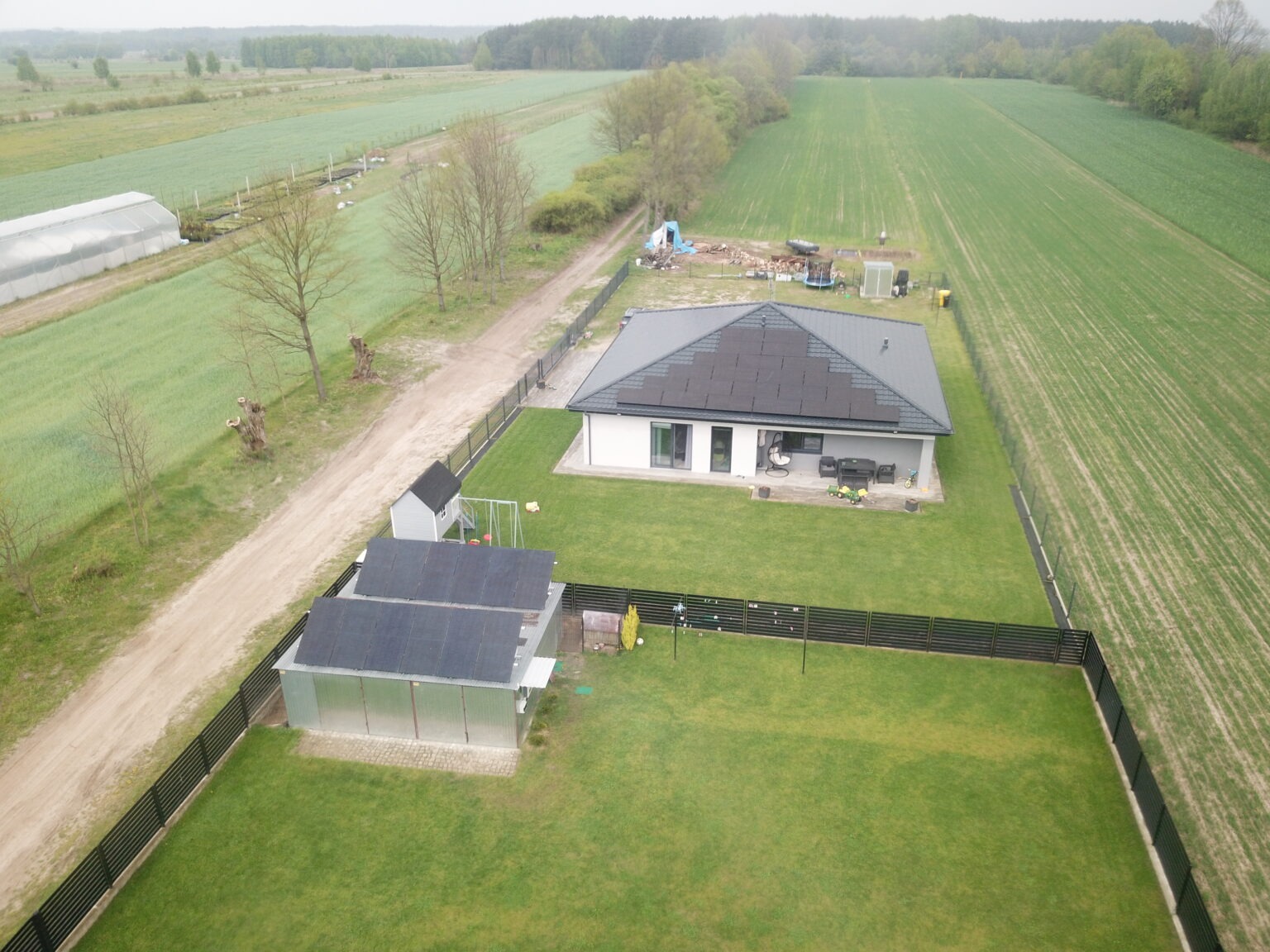 Widok z drona na dom jednorodzinny z panelami słonecznymi na dachu i na dachu budynku gospodarczego, otoczony zielonym trawnikiem i polami uprawnymi.