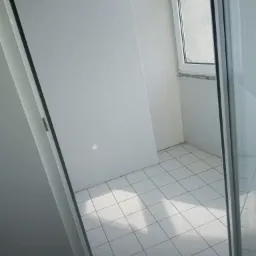 Wnętrze balkonu z białymi ścianami o lekko chropowatej strukturze, białą ramą okienną i białą podłogą wyłożoną kwadratowymi płytkami. Częściowo widoczne drzwi balkonowe.