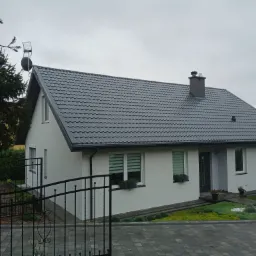 Jasny, jednorodzinny dom z szarą dach&oacute;wką, świeżo po remoncie elewacji, z anteną na dachu, widoczny zza metalowego ogrodzenia, z zadbanym ogrodem frontowym i fragmentem zaparkowanego samochodu.