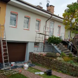 Dwukondygnacyjny dom jednorodzinny w trakcie prac elewacyjnych: część pomalowana na szaro, okna zabezpieczone folią, widoczne drabiny, rusztowanie, wiaderka z farbą i narzędzia malarskie...