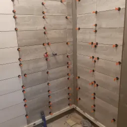 Ściana wyłożona płytkami imitującymi drewno, z użyciem systemu poziomowania z pomarańczowymi klinami, widoczne fragmenty instalacji hydraulicznej.