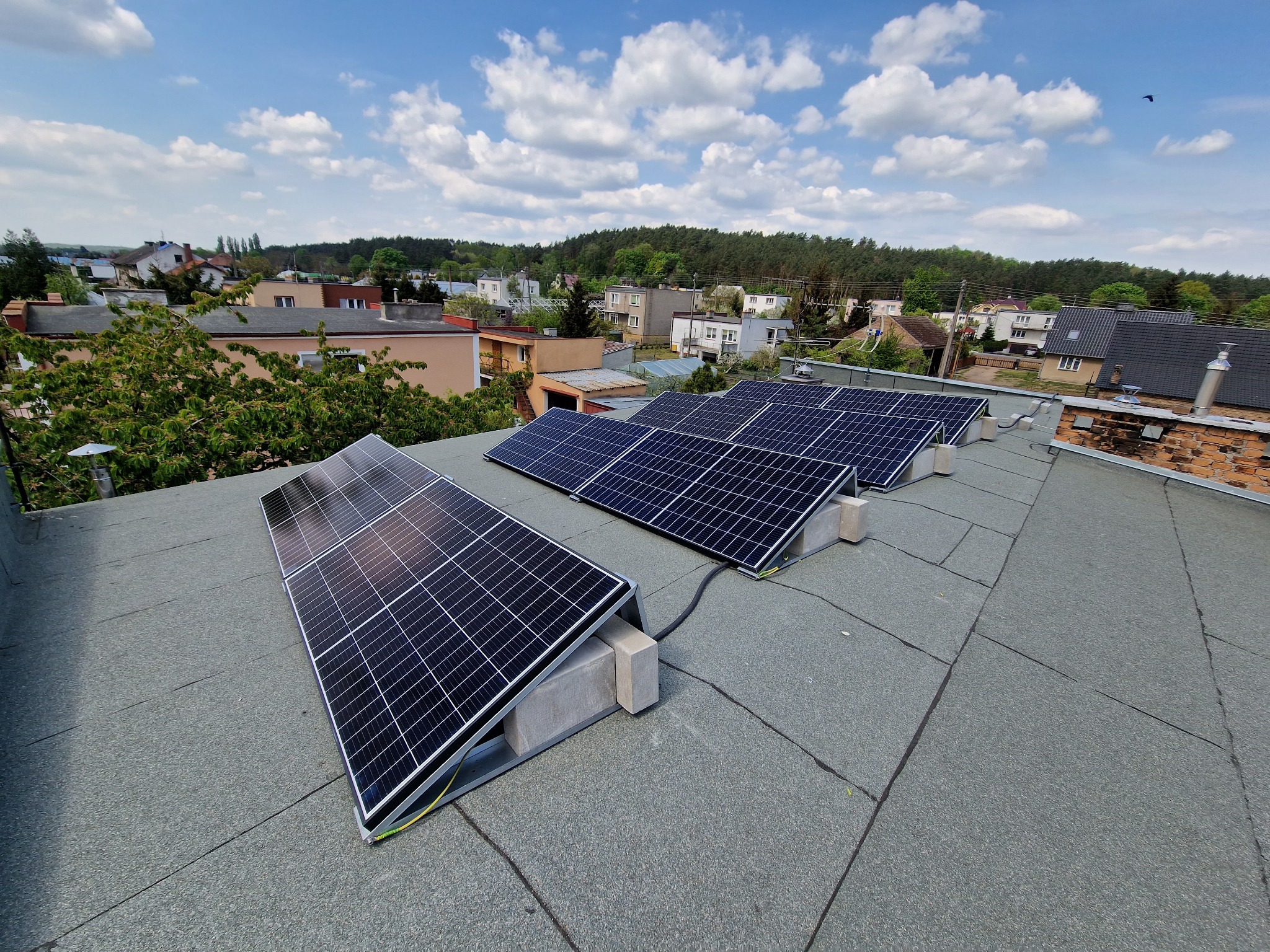 Panele słoneczne zamontowane na płaskim dachu budynku mieszkalnego, widok z góry na instalację z przewodami, w tle zabudowa jednorodzinna i błękitne niebo z chmurami.