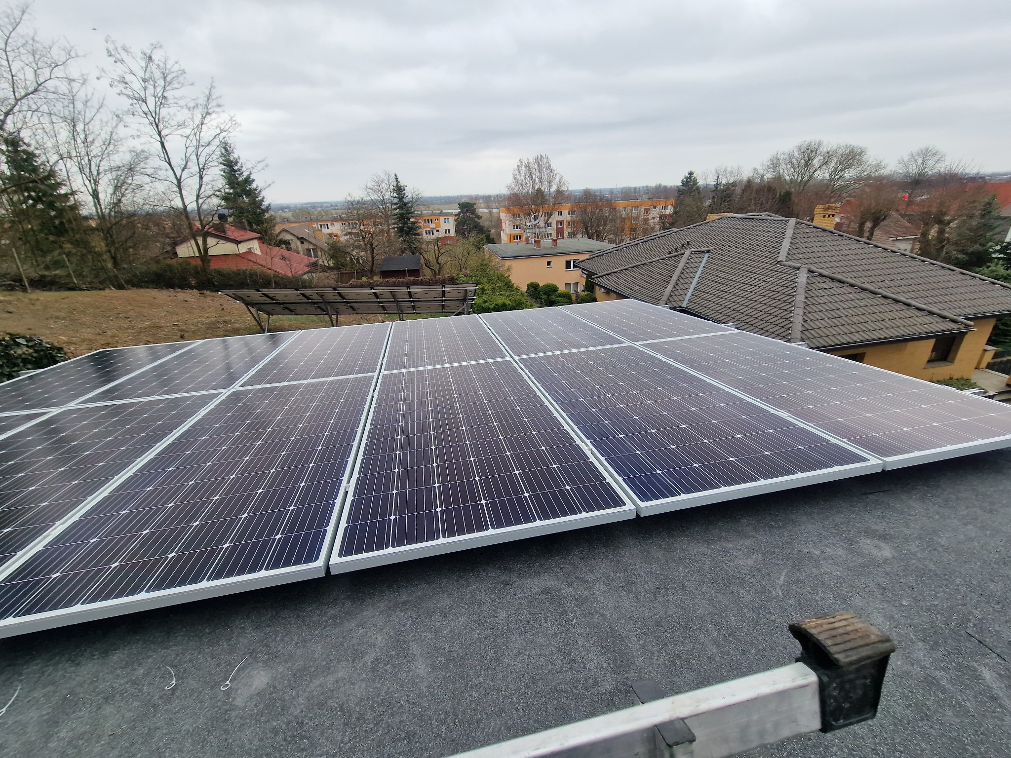 Panel słoneczny zainstalowany na dachu z widokiem na zabudowania mieszkalne w tle, drabina oparta o krawędź dachu.