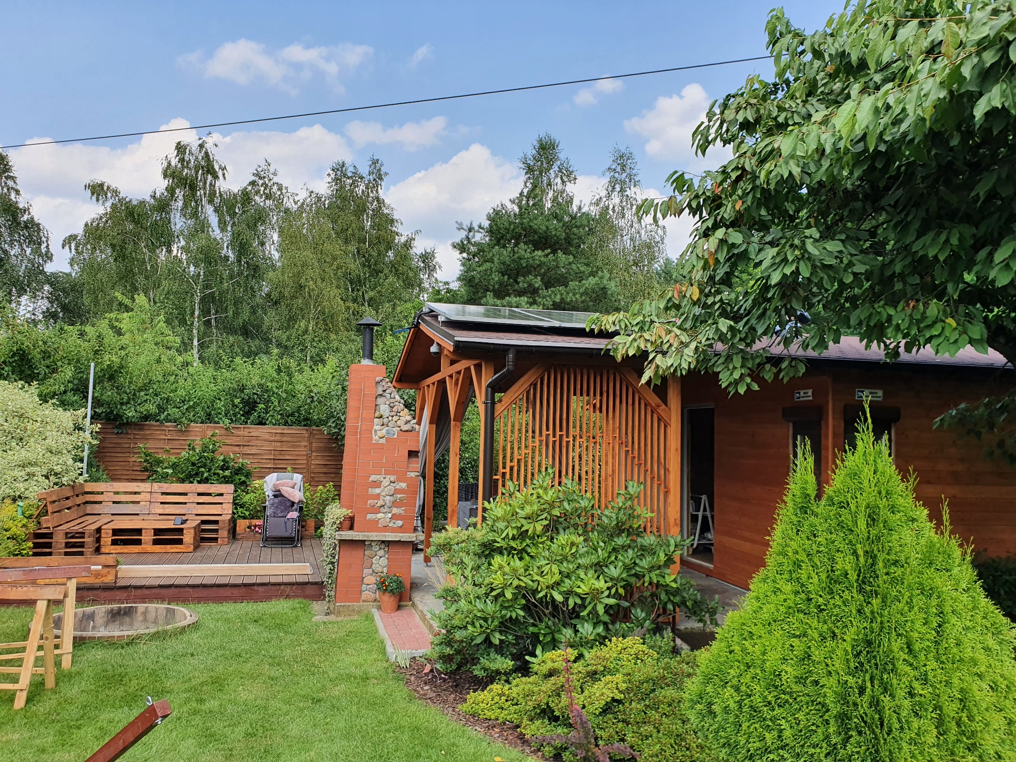 Instalacja paneli słonecznych na dachu drewnianej altany w Gorzowie Wielkopolskim, widoczna z poziomu trawnika, otoczona zielenią i drzewami.