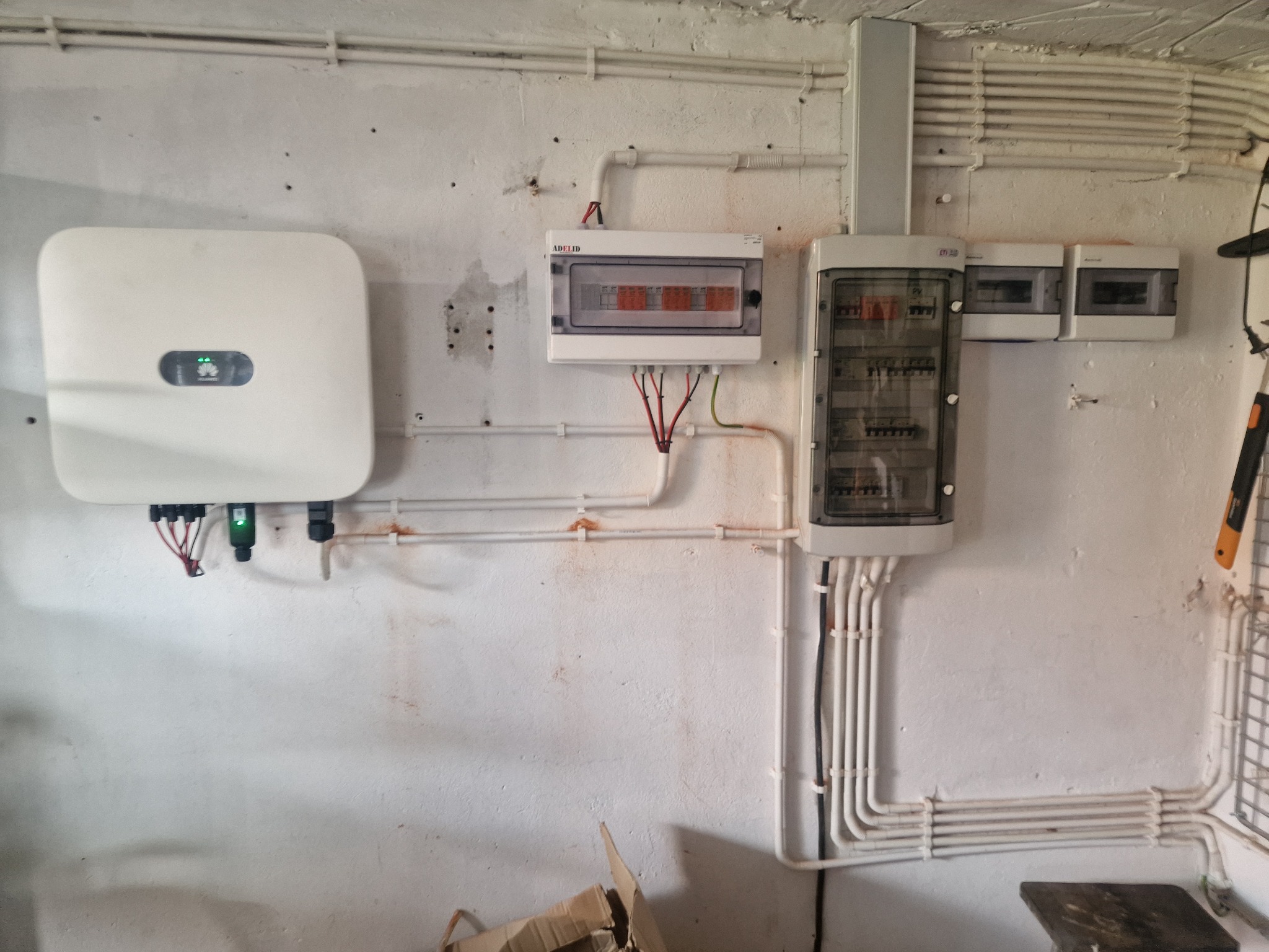 Instalacja fotowoltaiczna wewnątrz budynku: biały falownik Huawei z zielonymi diodami, rozdzielnice elektryczne marki Adelid i inne, białe rury instalacyjne na pomalowanej na biało ścianie...