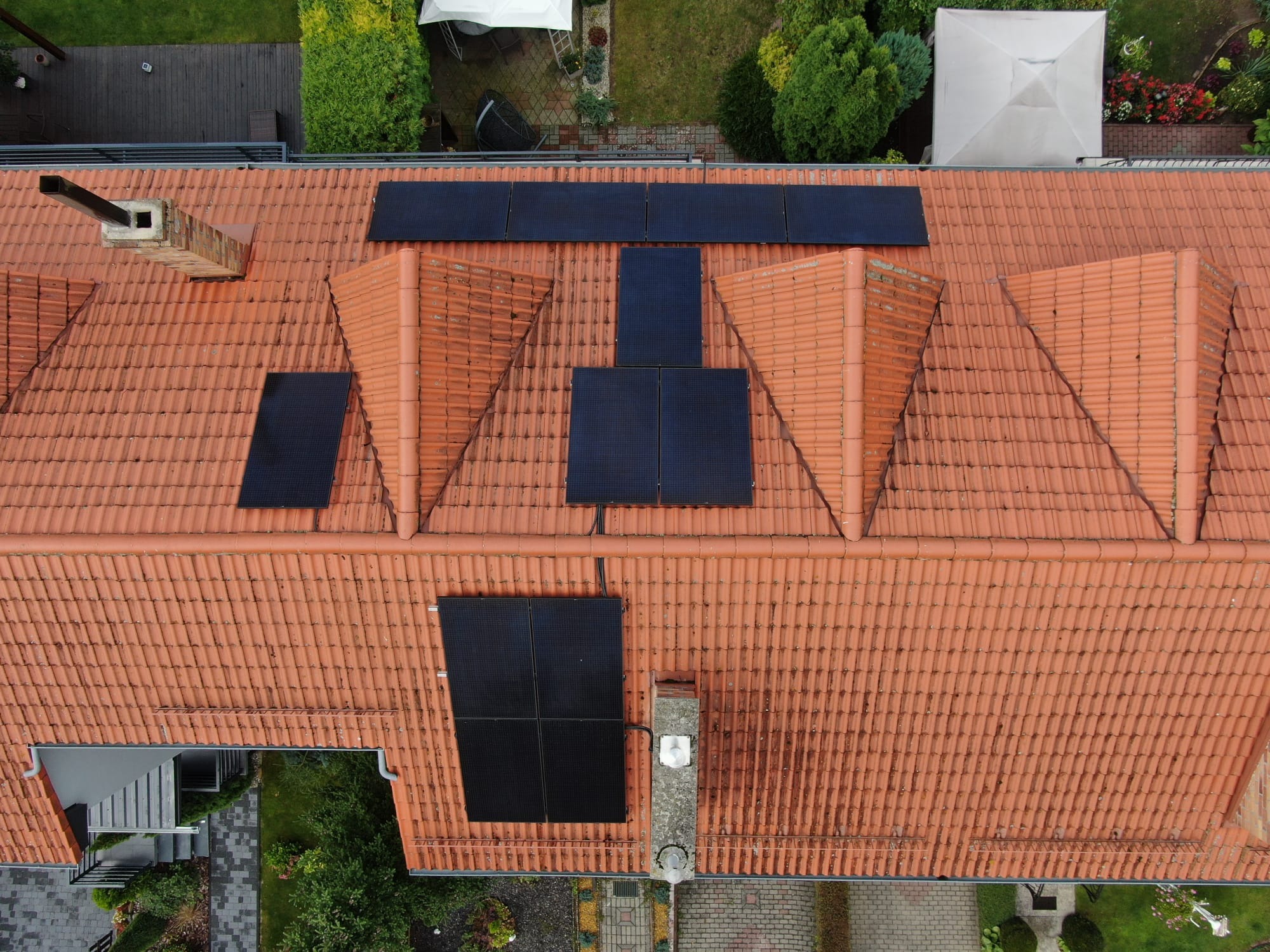 Widok z góry na dach z czerwonej dachówki z zamontowanymi panelami słonecznymi w układzie wschód-zachód, otoczony zielenią drzew i trawników.