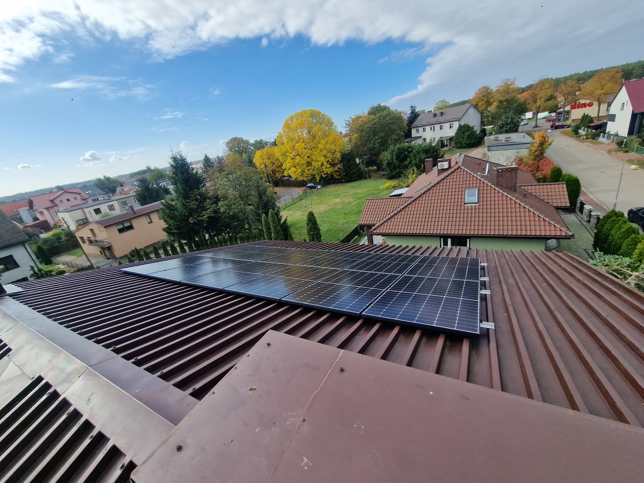 Panele słoneczne zamontowane na brązowym dachu blaszanym, widok z góry na okoliczne domy i drzewa w jesiennych barwach pod błękitnym niebem.