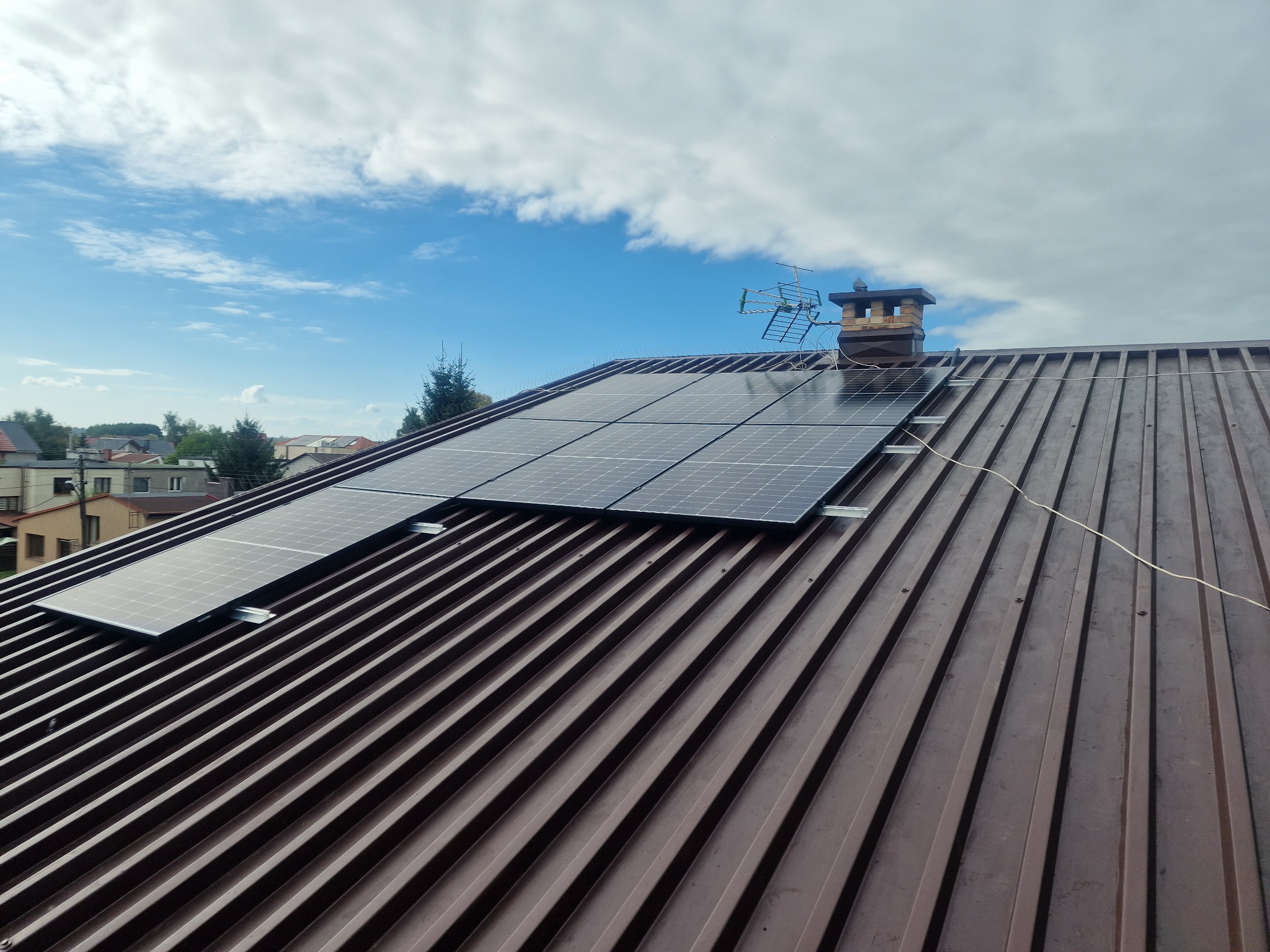 Panele słoneczne zamontowane na brązowym dachu z blachy trapezowej, widoczny komin z anteną telewizyjną na tle błękitnego nieba z chmurami.