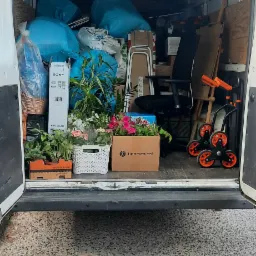 Wnętrze białego busa załadowane do przeprowadzki: niebieskie worki, kartony, kwiaty w doniczkach, biurowe krzesło i wózek transportowy z pomarańczowymi kołami.