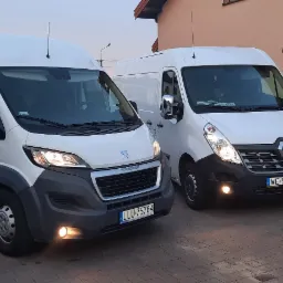 Dwa białe busy, Peugeot Boxer i Renault Master, zaparkowane obok siebie na brukowanym podjeździe przed domem z czerwonym dachem, światła włączone.