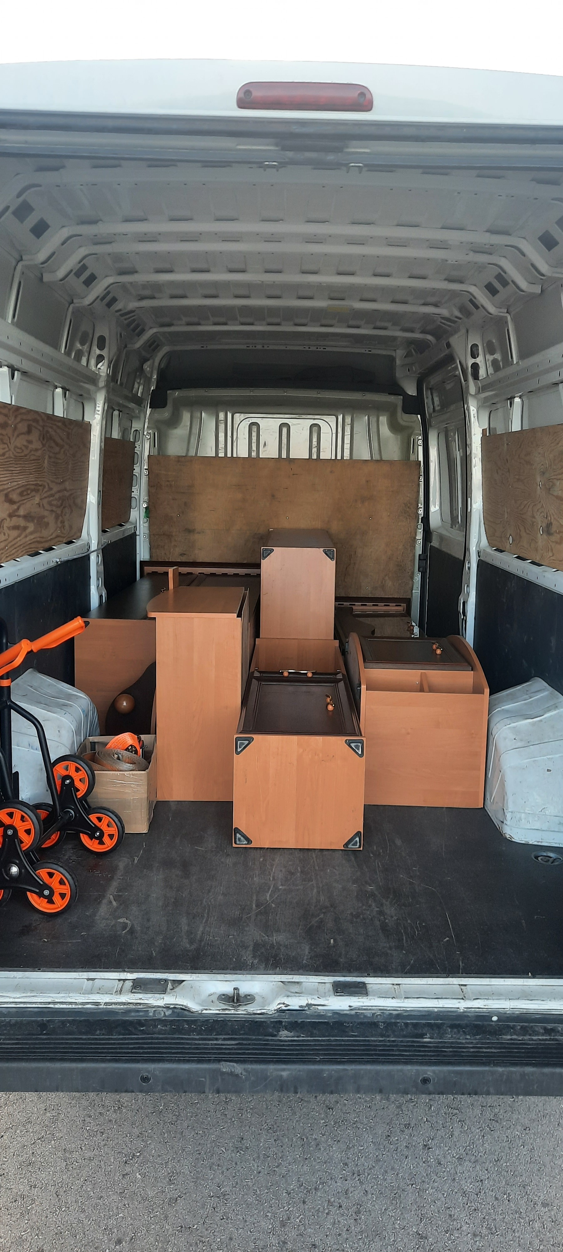 Wnętrze dostawczego busa wypełnione meblami z jasnego drewna, zabezpieczonymi na czas transportu, z widocznym wózkiem transportowym z pomarańczowymi kółkami.