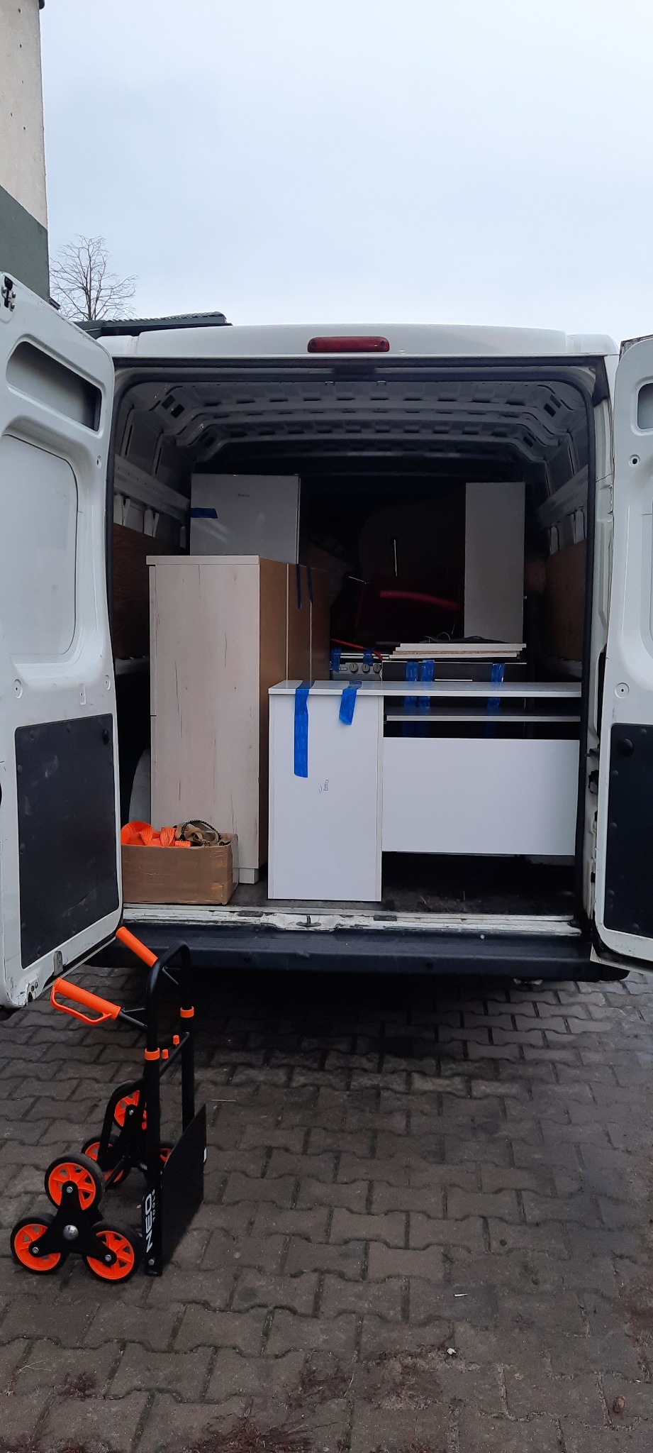 Wnętrze białego busa z otwartymi drzwiami załadowane meblami zabezpieczonymi niebieską taśmą, na zewnątrz wózek transportowy NEO z pomarańczowymi kołami.