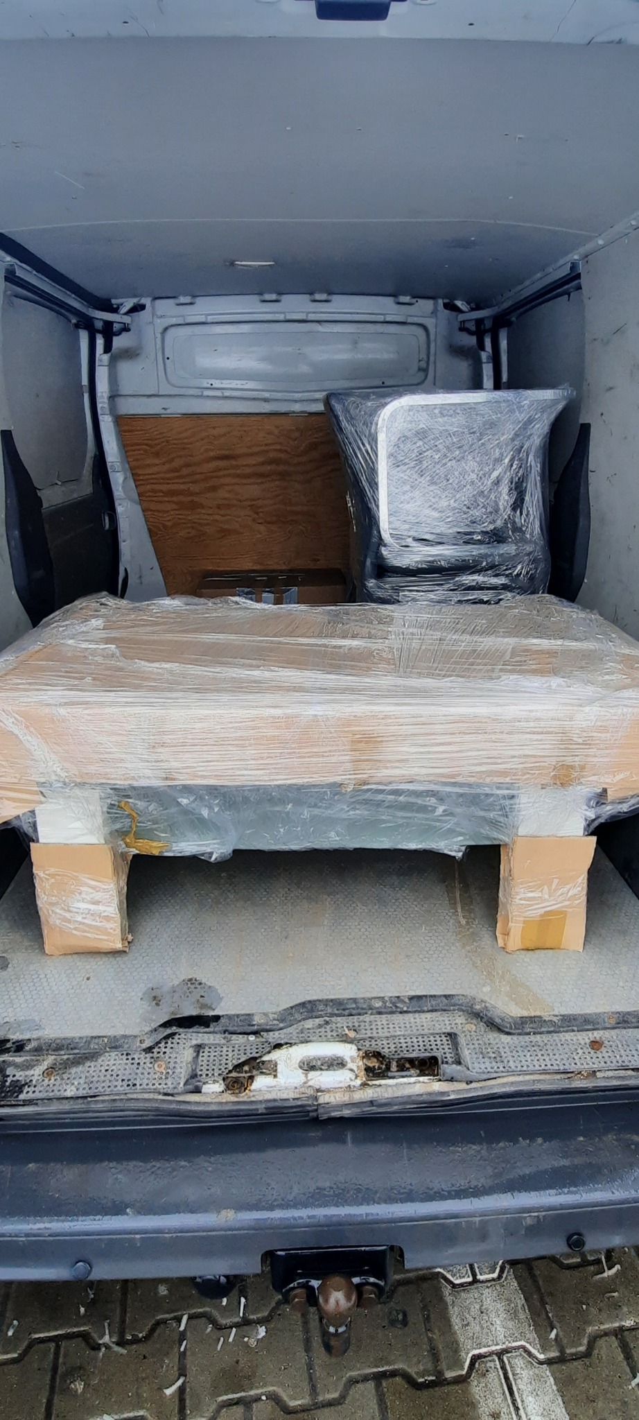 Wnętrze furgonetki z zabezpieczonymi folią i kartonami meblami, gotowymi do transportu. Widoczny stary fotel biurowy owinięty folią stretch.