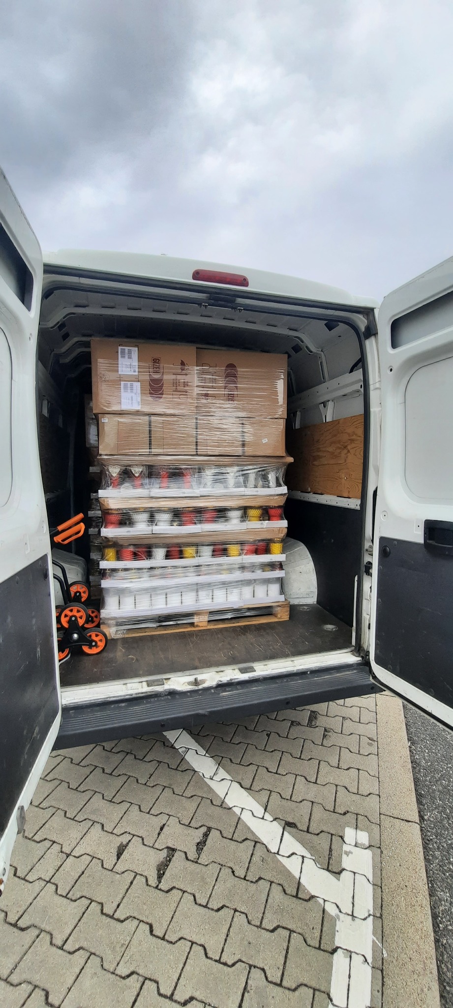 Wnętrze białego busa z otwartymi drzwiami załadowane paletą z zapakowanymi kartonami i przezroczystą folią towarami, dodatkowo z lewej strony widać wózek transportowy z pomarańczowymi kółkami.