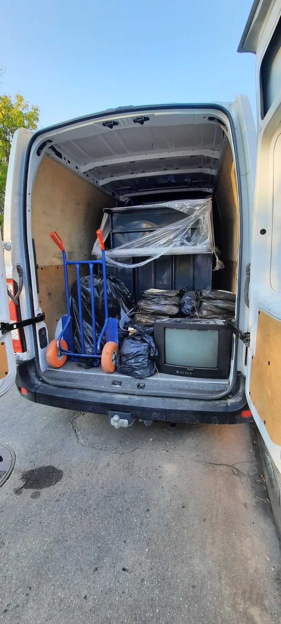 Wnętrze białego busa z otwartymi drzwiami załadowane meblami, workami z rzeczami i starym telewizorem Philips, z wózkiem transportowym z pomarańczowymi kołami z boku.