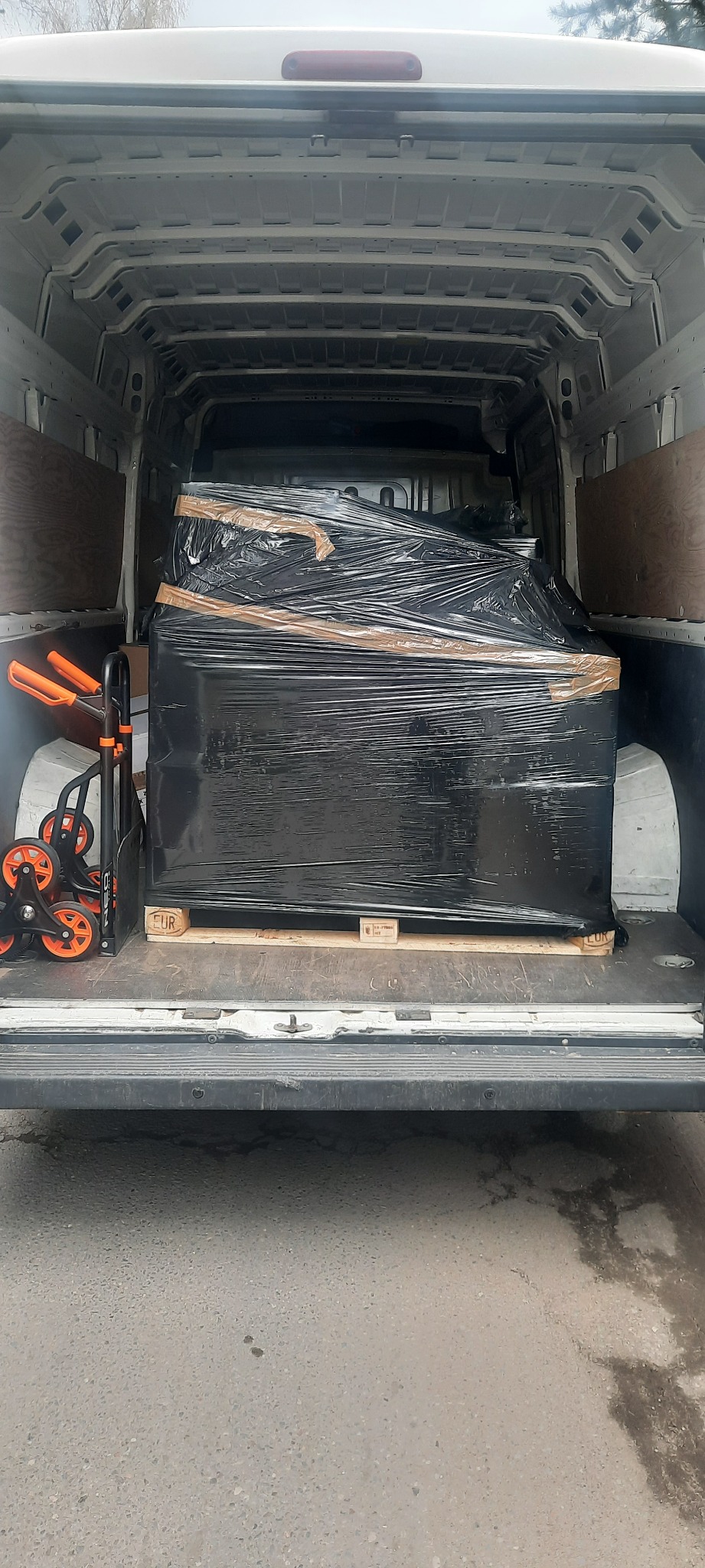 Wnętrze białego furgonetki z ładunkiem owiniętym czarną folią na palecie EUR, obok wózek transportowy z pomarańczowymi kółkami.