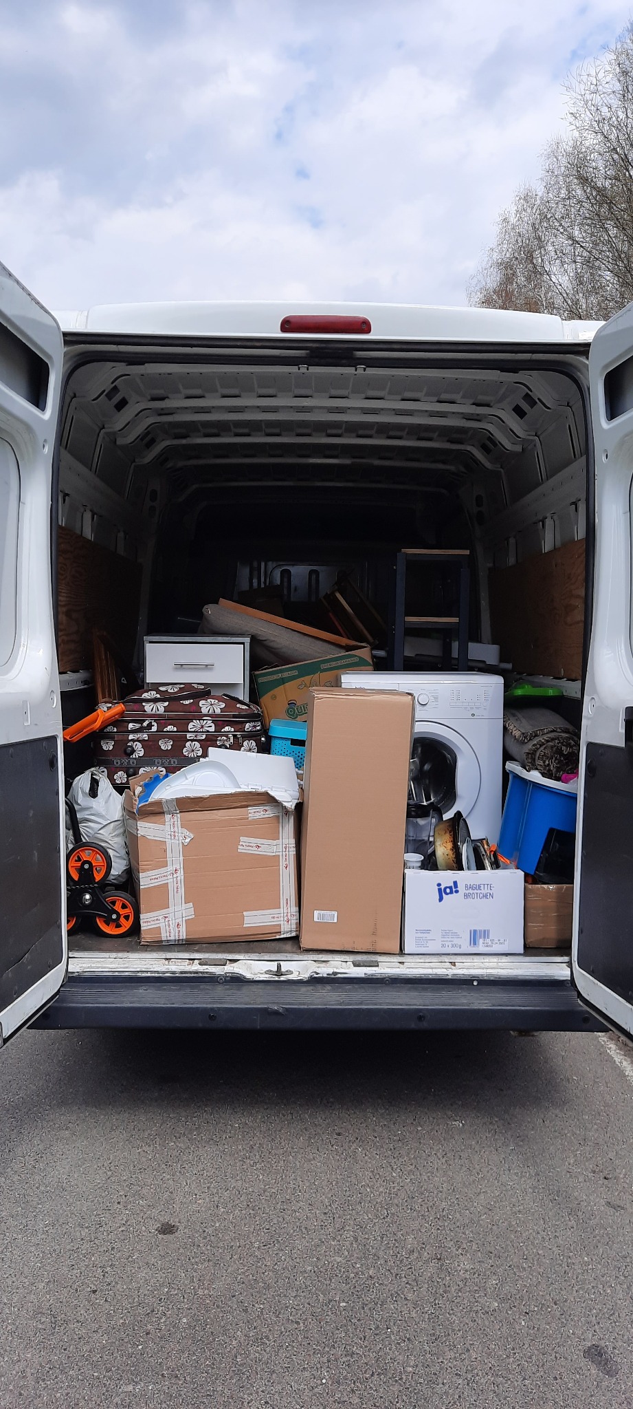 Wnętrze białego busa załadowane kartonami, meblami i pralką, widok z tyłu przez otwarte drzwi.