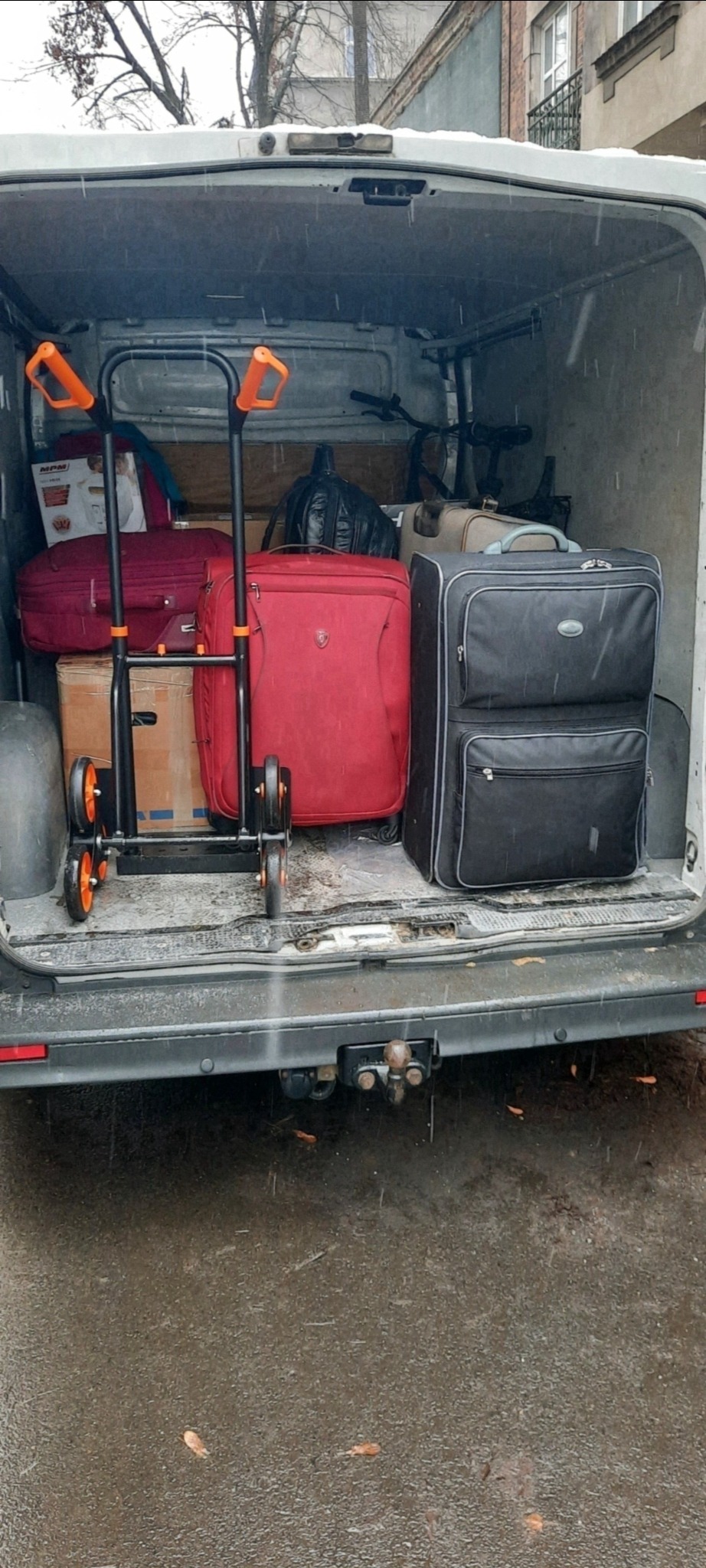 Wnętrze brudnego busa załadowane bagażami, w tym walizkami, torbami i kartonami, oraz wózkiem transportowym z pomarańczowymi uchwytami, widoczne w deszczowy dzień.