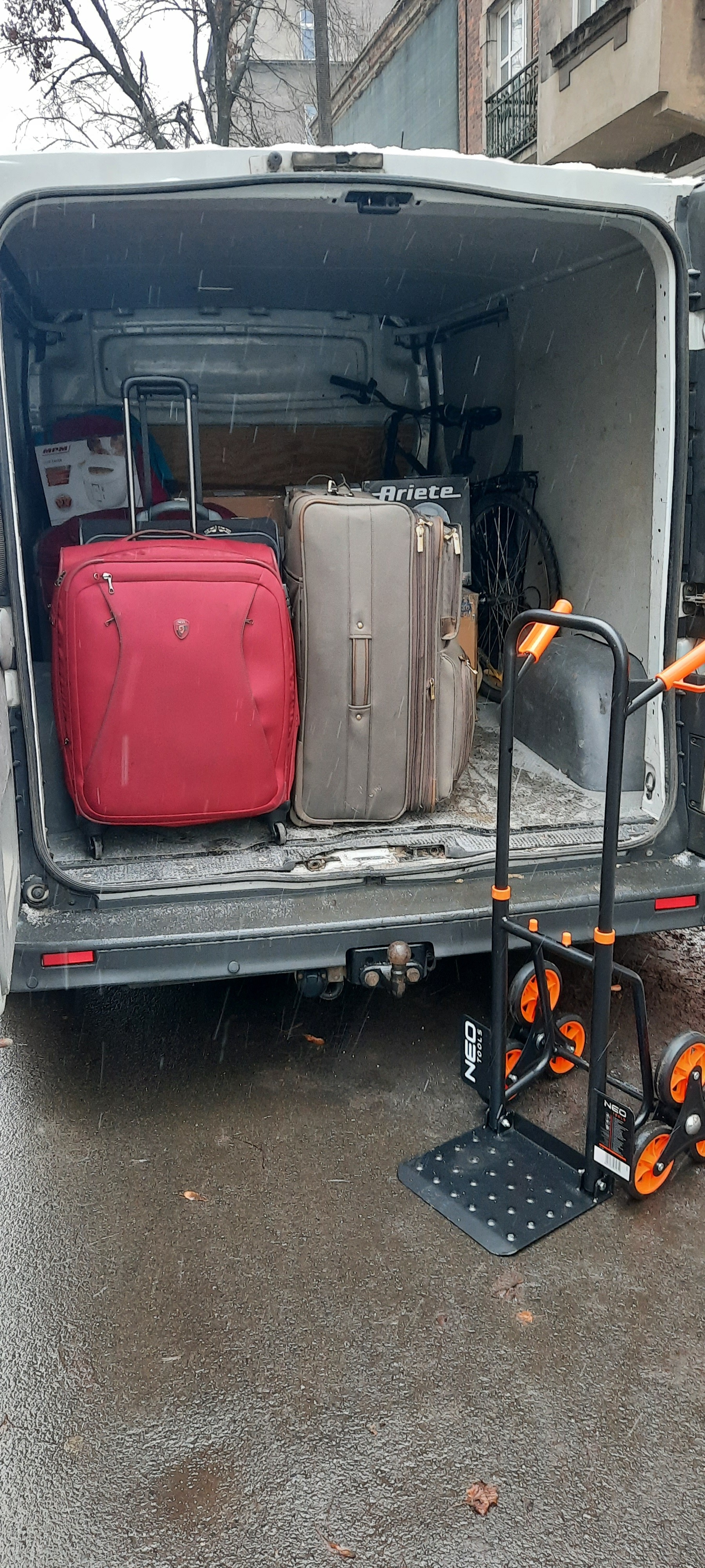 Otwarty bagażnik busa załadowany walizkami, kartonami i rowerem, obok wózek transportowy Neo Tools na mokrym asfalcie w deszczowy dzień.