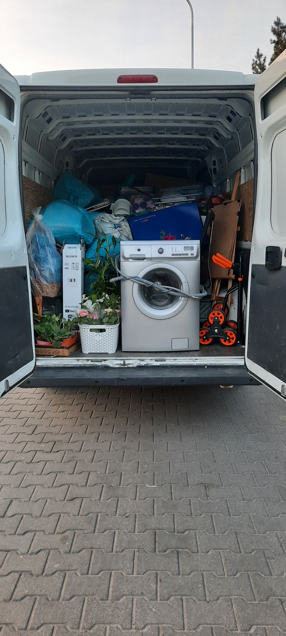 Wnętrze białego furgonetki z otwartymi drzwiami, załadowane sprzętem AGD, roślinami doniczkowymi i workami z rzeczami, gotowe do transportu.