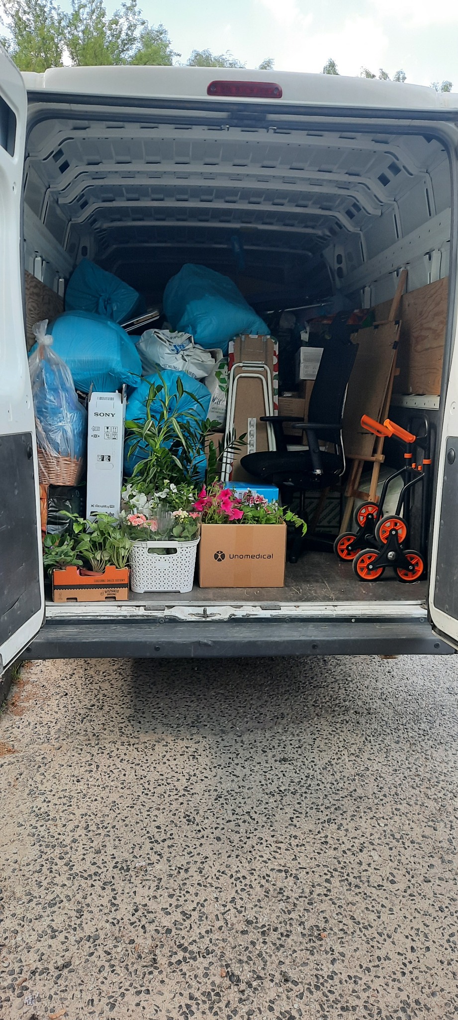 Wnętrze białego busa załadowane do przeprowadzki: niebieskie worki, kartony, kwiaty w doniczkach, biurowe krzesło i wózek transportowy z pomarańczowymi kołami.