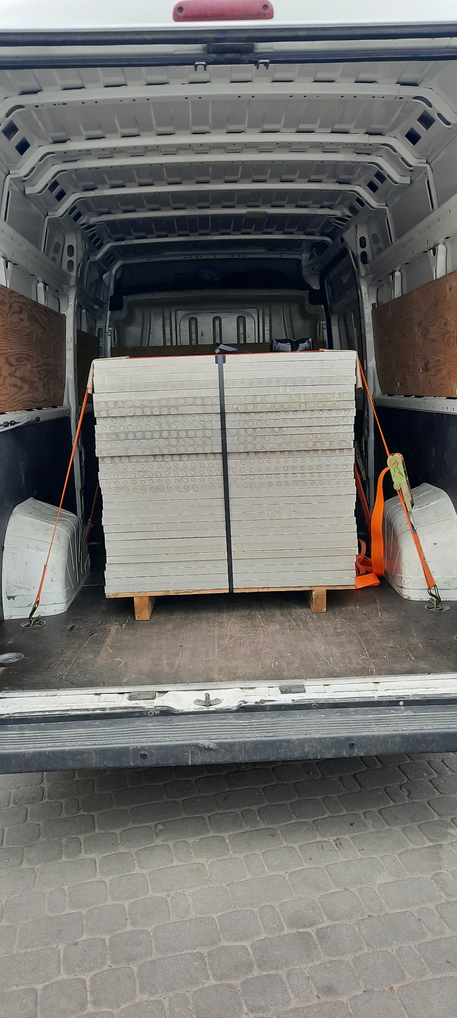 Paleta płyt z otworami zabezpieczona pasami w przestrzeni ładunkowej białego busa, widok z tyłu.