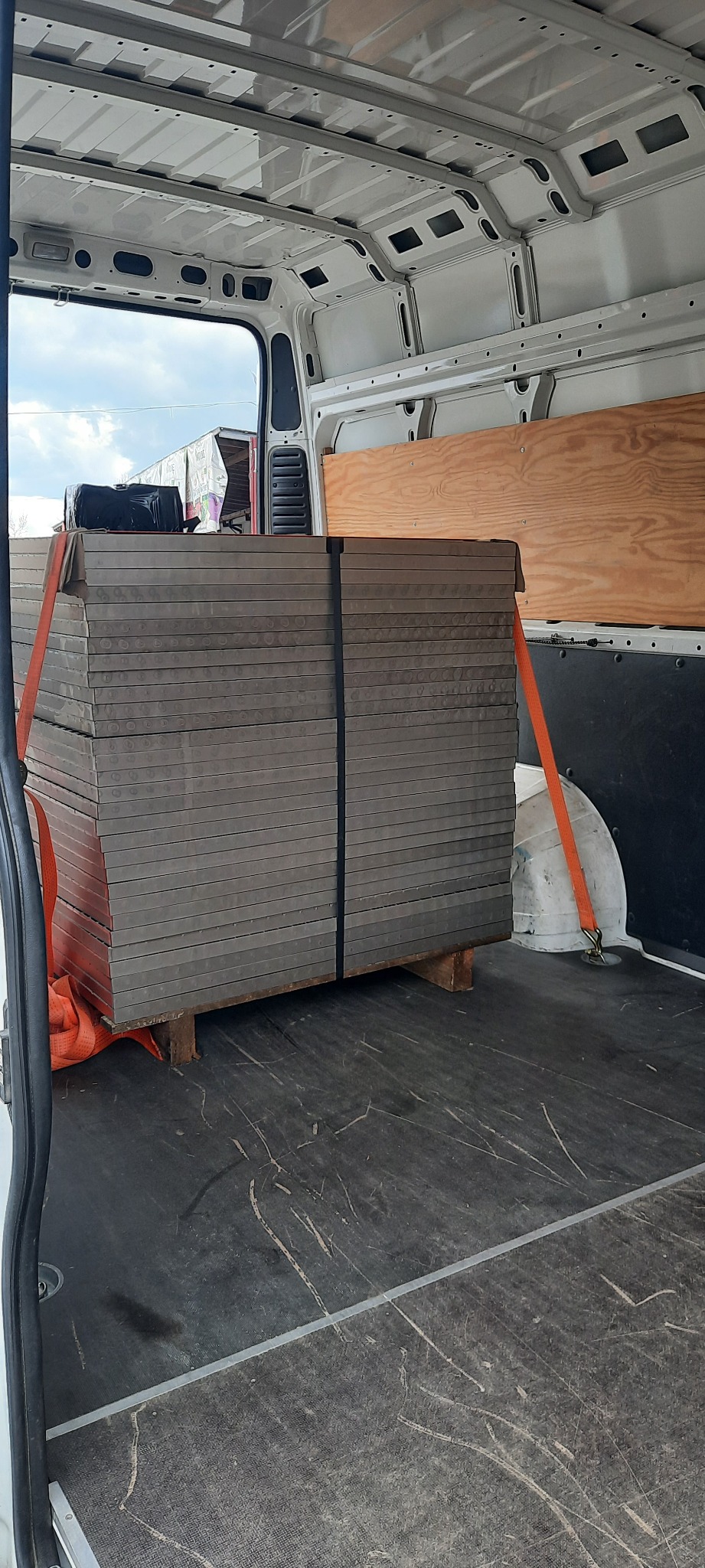 Wnętrze białego busa z załadowanym towarem zabezpieczonym pomarańczowymi pasami transportowymi, widoczne palety i czarna torba na górze.