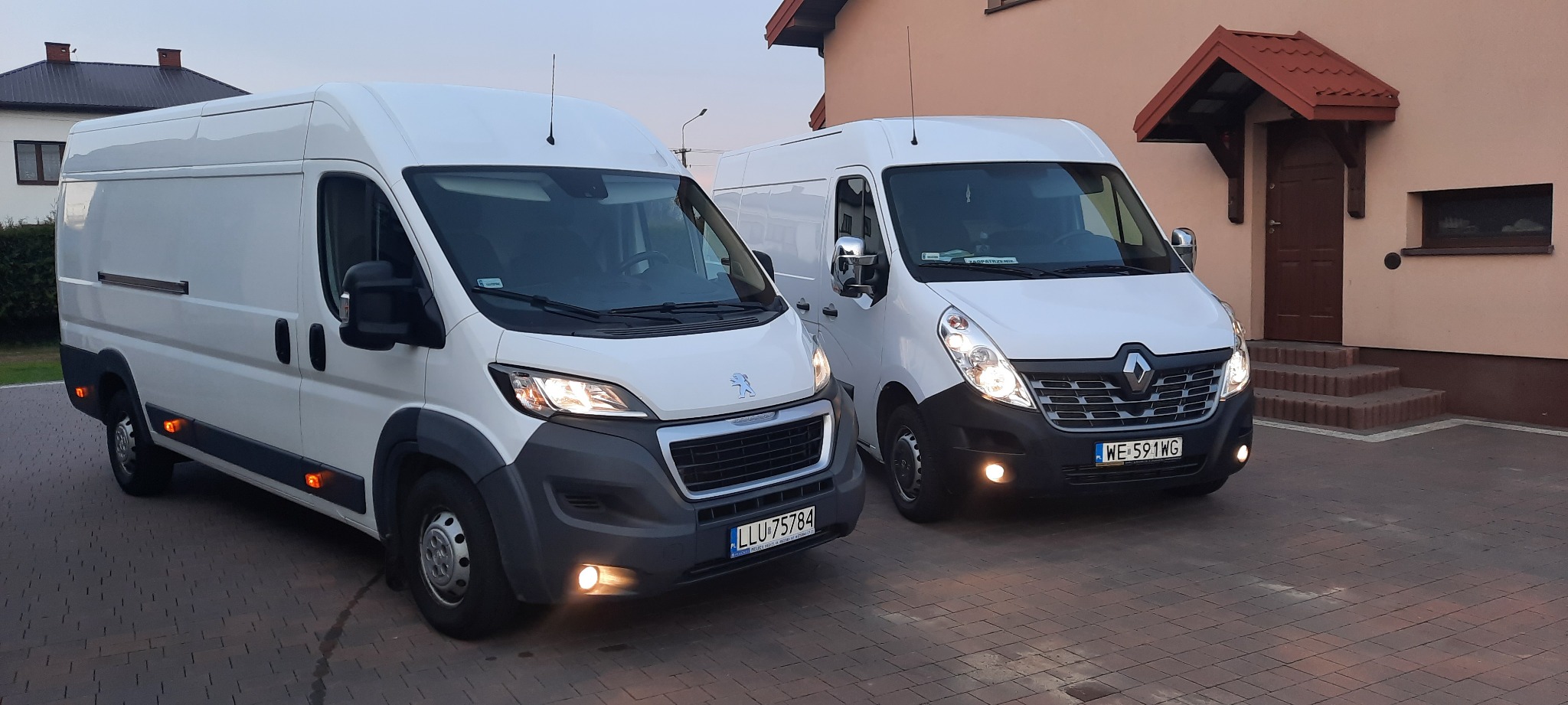 Dwa białe busy, Peugeot Boxer i Renault Master, zaparkowane obok siebie na brukowanym podjeździe przed domem z czerwonym dachem, światła włączone.