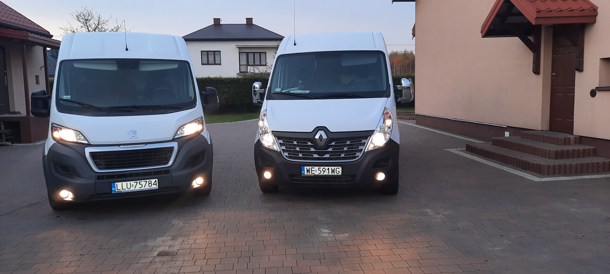 Dwa białe busy, Peugeot i Renault Master, zaparkowane obok siebie na brukowanym podjeździe przed domem jednorodzinnym, z włączonymi światłami mijania i przeciwmgielnymi.