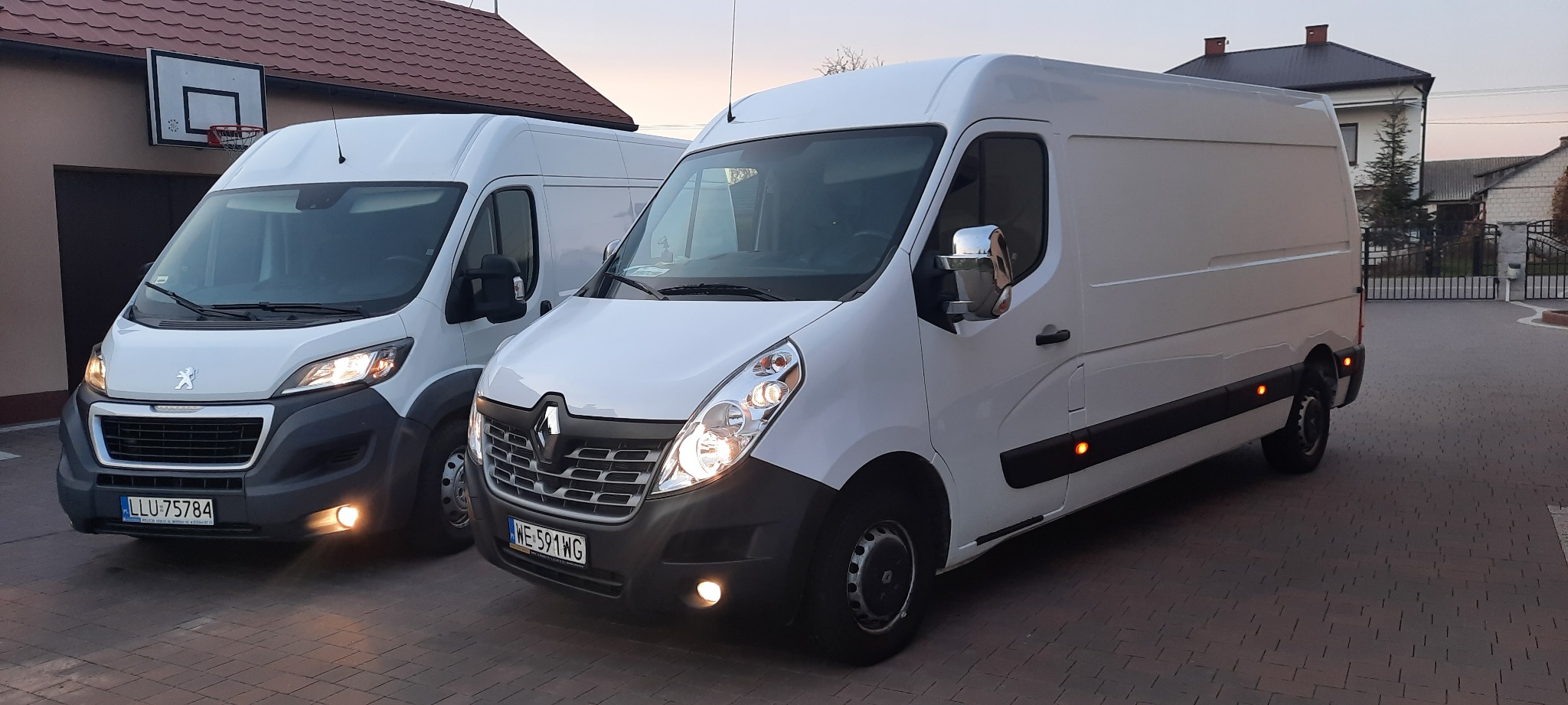 Dwa białe busy, Peugeot i Renault Master, zaparkowane na brukowanym podjeździe przed domem z koszem do koszykówki, światła włączone.