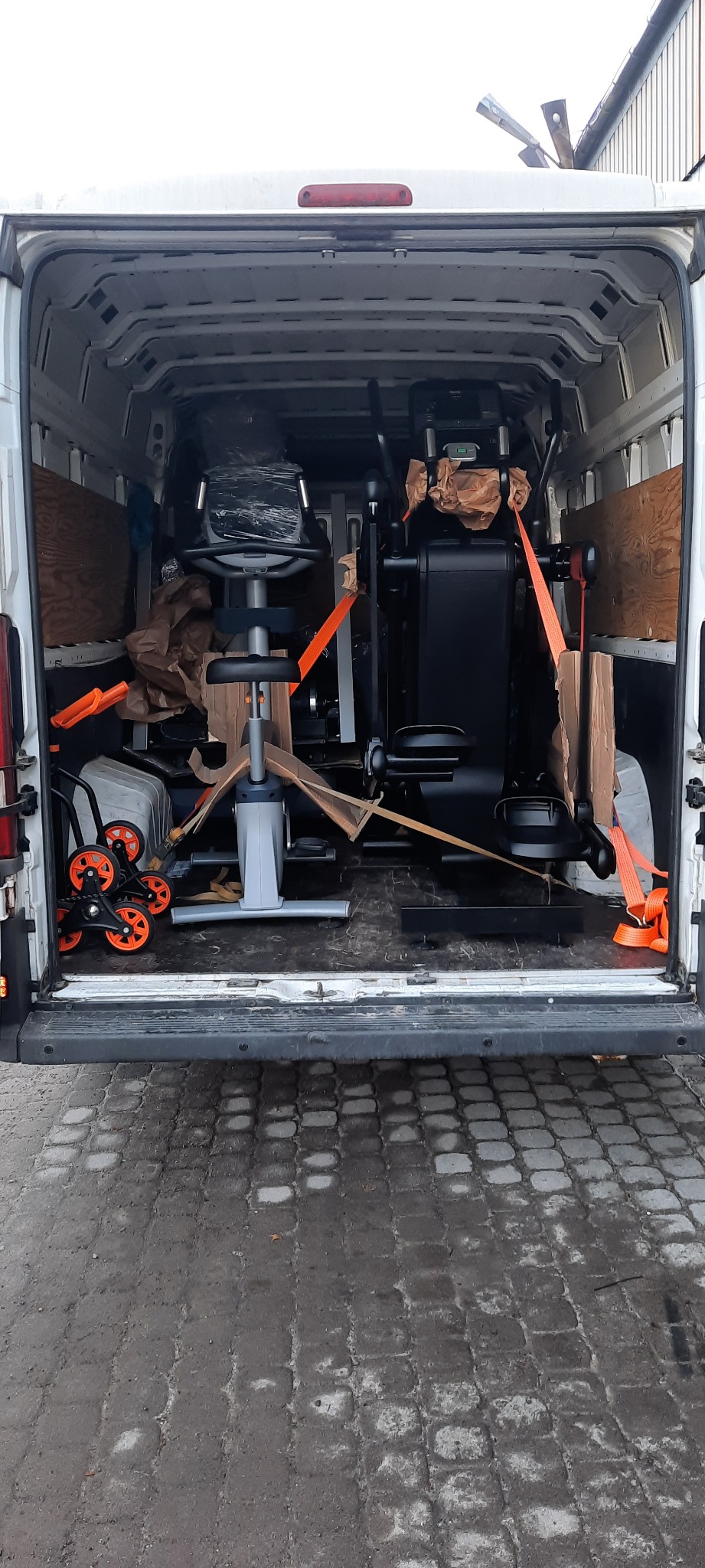 Wnętrze białego furgonetki z widocznymi maszynami do ćwiczeń, zabezpieczonymi pomarańczowymi pasami, przygotowanymi do transportu.