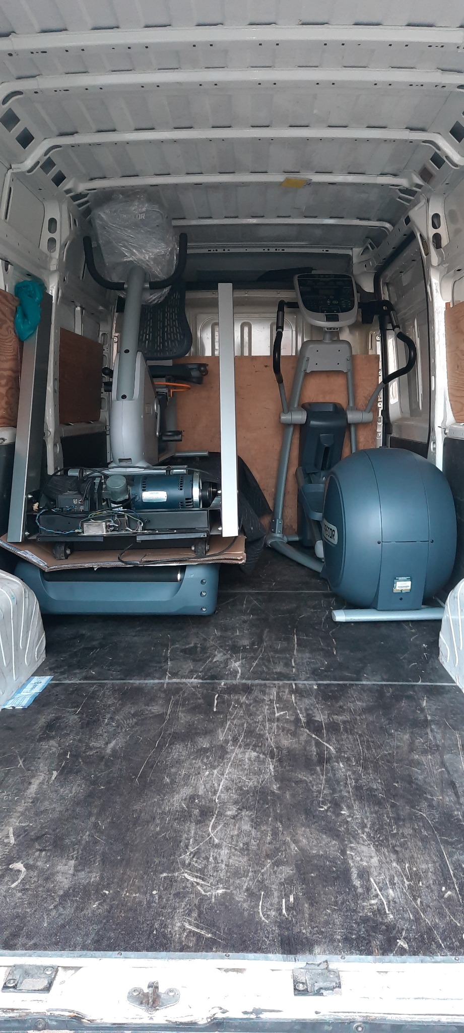 Wnętrze busa wypełnione sprzętem do ćwiczeń, w tym eliptykami i rozmontowanymi maszynami, częściowo zabezpieczonymi folią.