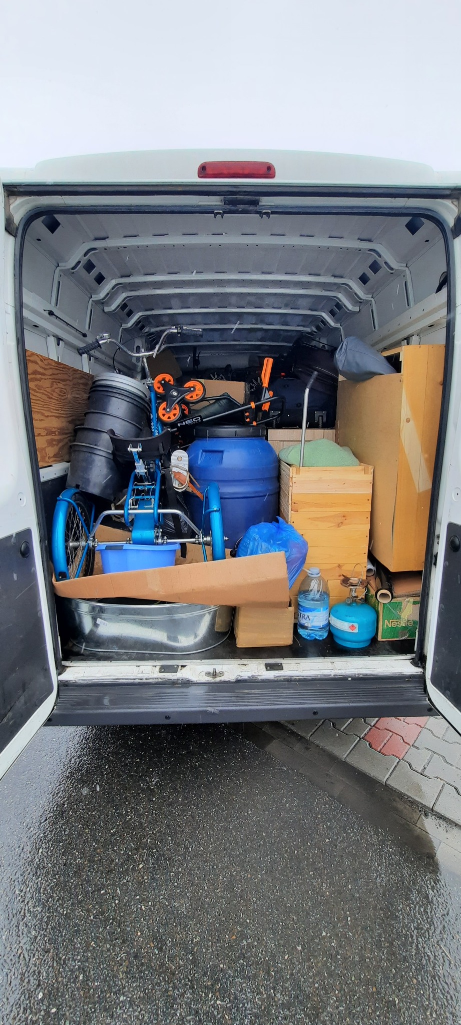 Wnętrze białego furgonetki załadowane różnymi przedmiotami, w tym niebieski trójkołowy rower, plastikowe beczki, drewniane skrzynie i butla gazowa, widoczne przez otwarte tylne drzwi na mokrym...