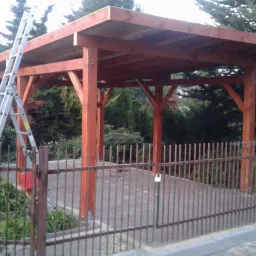 Drewniana wiata garażowa w trakcie budowy, widoczna drabina oparta o konstrukcję, metalowe ogrodzenie z przodu, dom w tle.