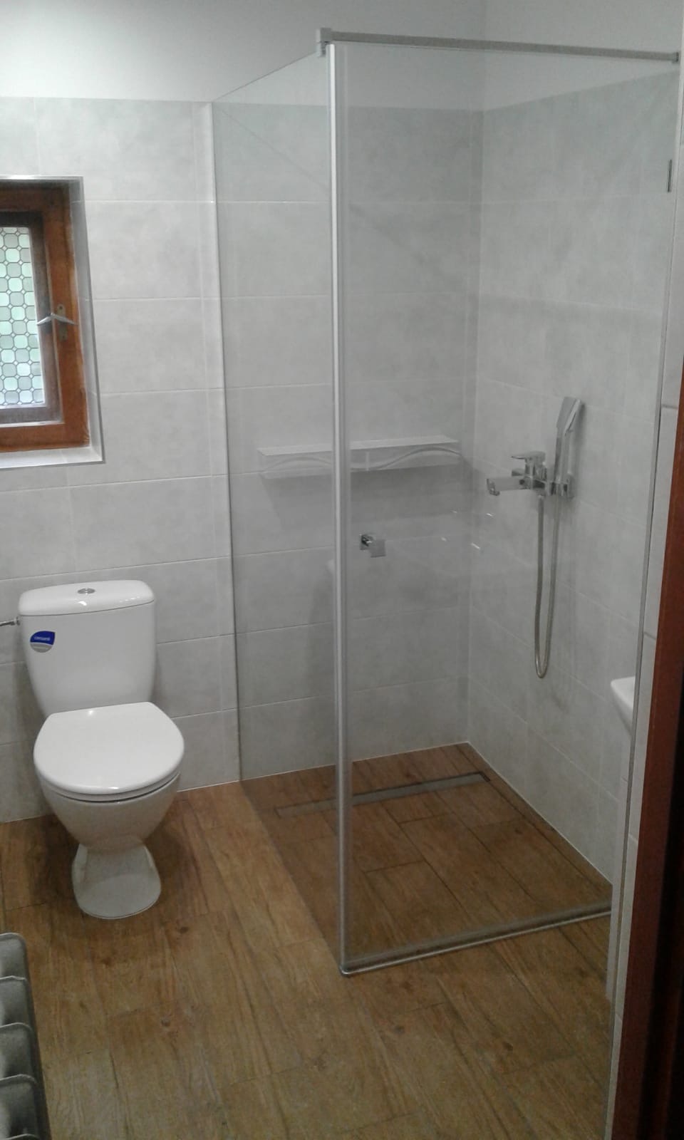 Nowoczesna łazienka z kabiną prysznicową ze szklaną ścianką, toaleta kompaktowa i drewnopodobne płytki na podłodze, okno z ozdobną szybą.